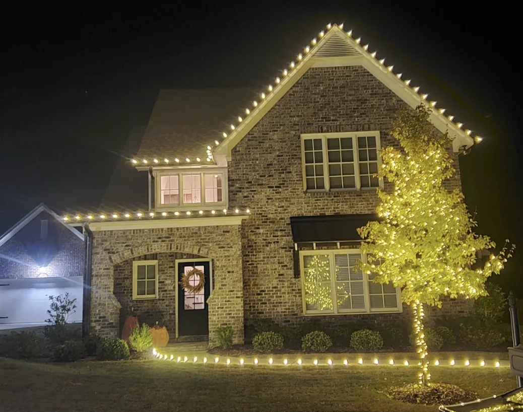 Brick home outlined in warm white lights with a lit tree in the front yard